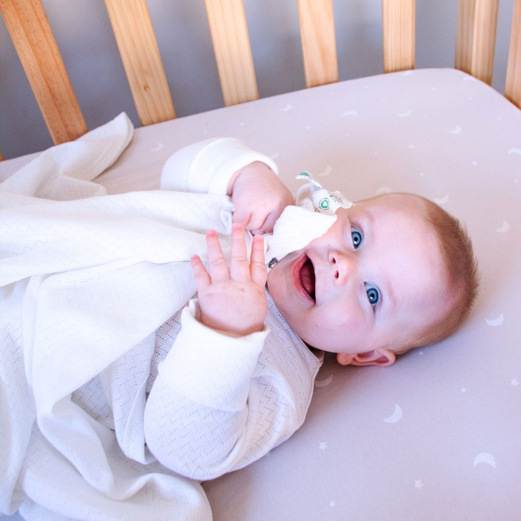 Baby lying in a crib with a white blanket, looking surprised or excited.