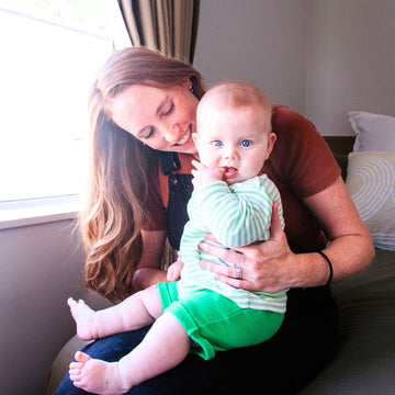 Woman holding a baby in a room with a window and pillows.