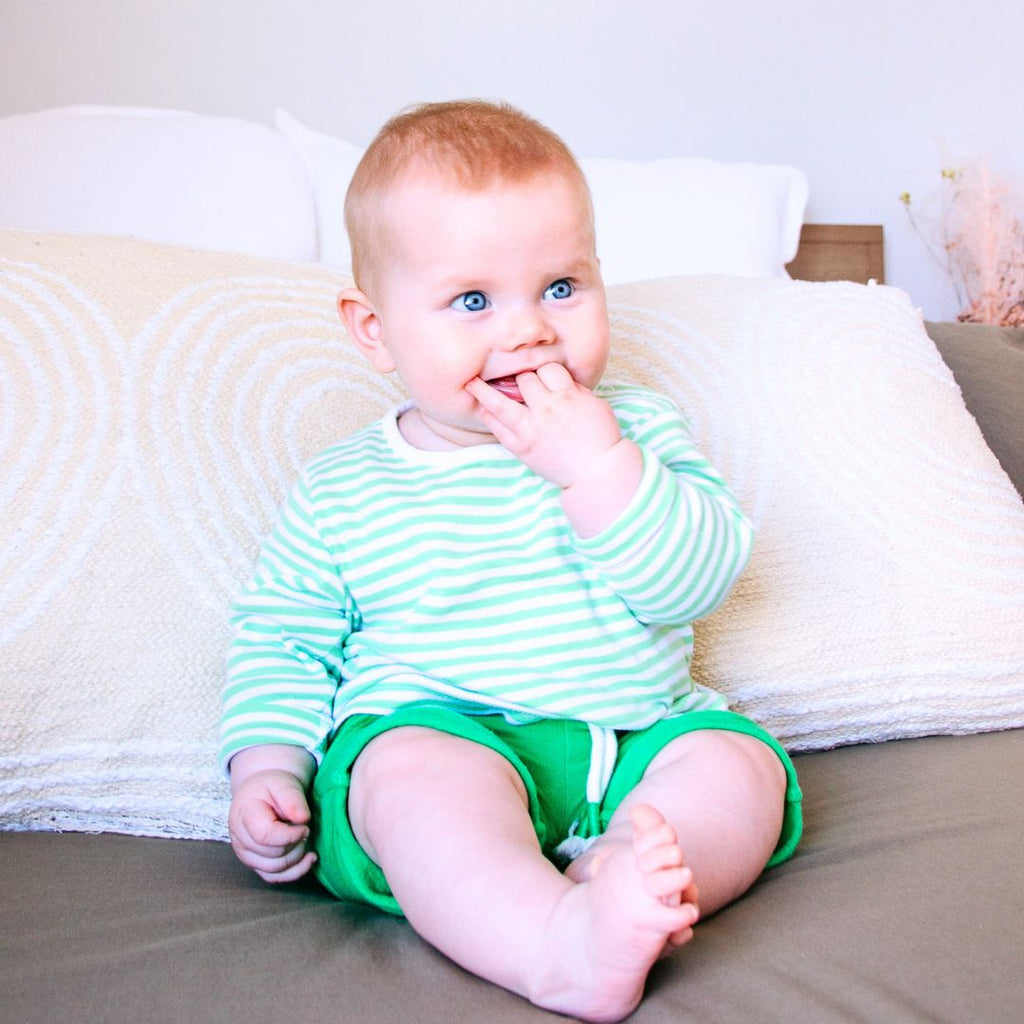 Baby in a striped shirt and green shorts sitting on a bed.