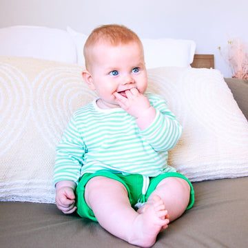 Baby in a striped shirt and green shorts sitting on a bed.