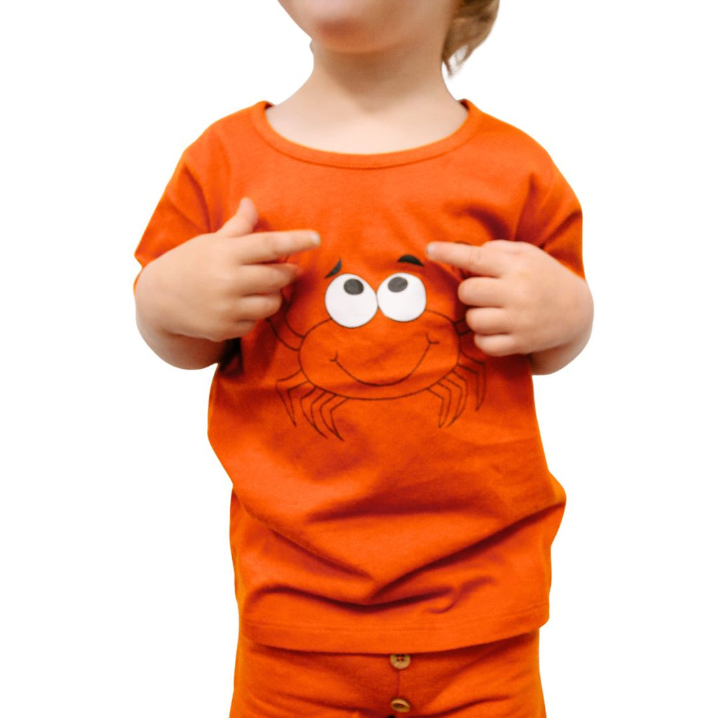 Child wearing an orange tee shirt with a crab design, pointing at it.