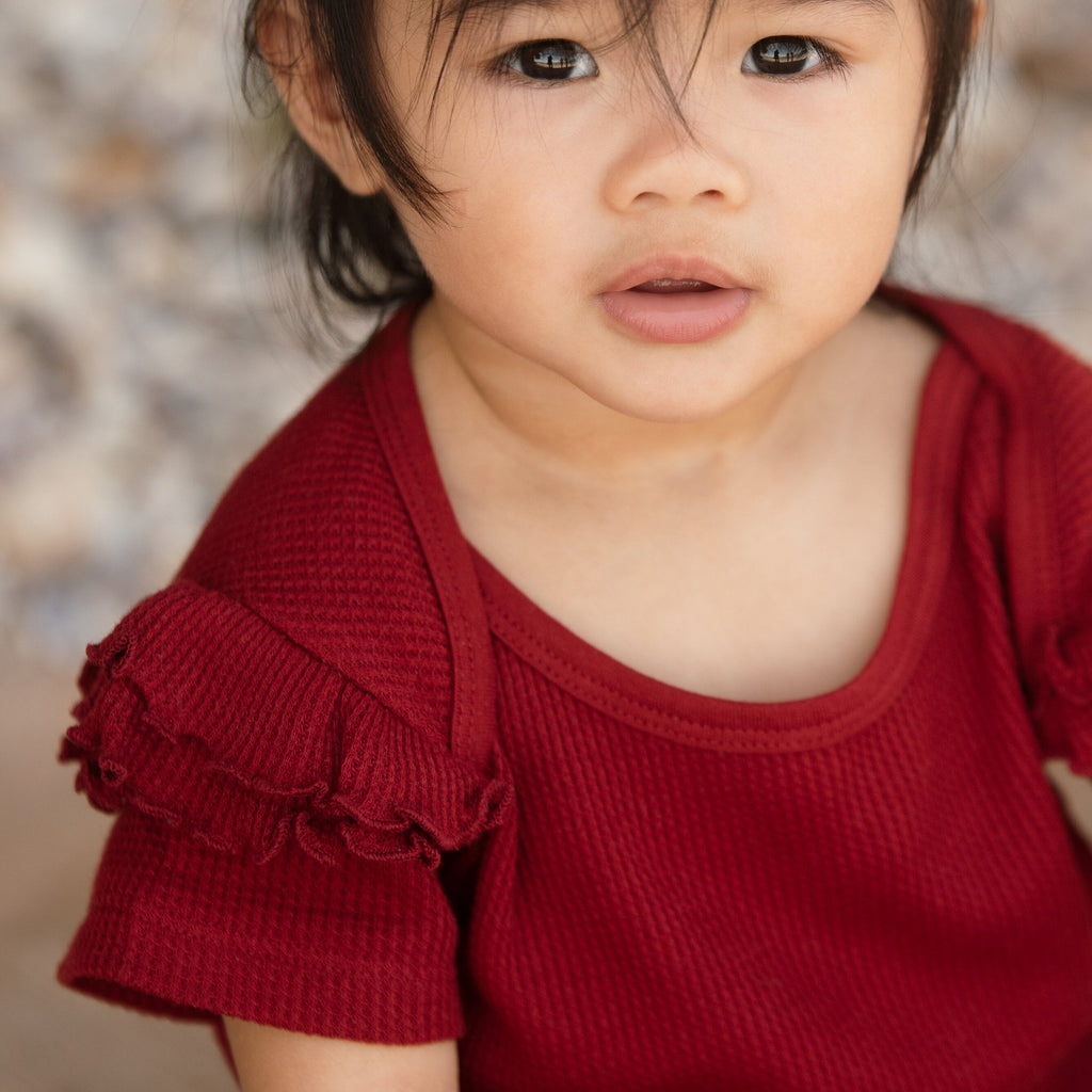 A close-up image of a toddler wearing a red organic cotton wine ruffled bodysuit with short puffed sleeves.